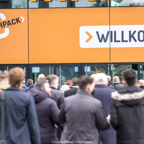 Man sieht eine geschäftige Menschenmenge, die einen Ausstellungsbereich betritt. Über dem Eingang stehen große orangefarbene Banner mit der Aufschrift „FACHPACK“ und „WILLKOMMEN“, die die Besucher auf Deutsch willkommen heißen. Die Atmosphäre, wahrscheinlich für die Fachpack 2024, ist lebhaft und voller Spannung und Spannung liegt in der Luft.