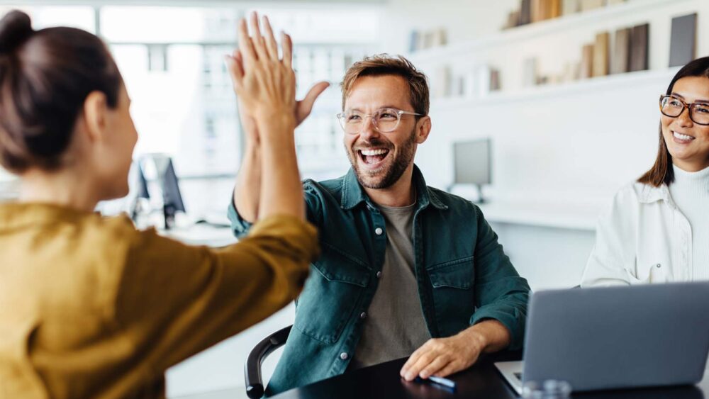 Drei Personen sitzen um einen Tisch in einem Büro. Ein Mann in der Mitte, der eine Brille und ein grünes Hemd trägt, lächelt breit, während er einer Frau links ein High Five gibt. Eine andere Frau sitzt lächelnd rechts mit einem Laptop vor sich und einer Präsetnationsverpackung daneben.