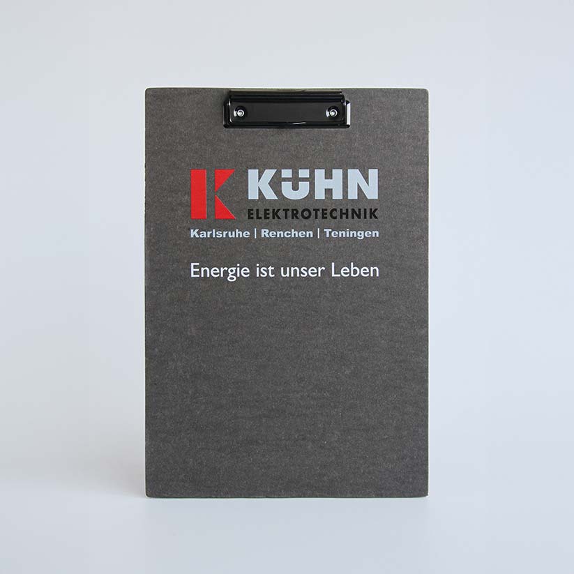 Ein schwarzes Klemmbrett mit einem Metallclip oben, auf dem das Logo und der Name „KÜHN ELEKTROTECHNIK“ in weißen und roten Buchstaben zu sehen sind, was moderne Drucktechniken zeigt. Darunter stehen die Worte „Karlsruhe | Renchen | Teningen“ und „Energie ist unser Leben“ in weißer Schrift.